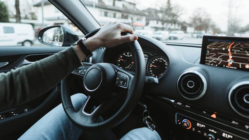 Arm steuert ein Auto, während GPS-Navigation auf dem Bildschirm angezeigt wird. Außen sind Fahrzeuge und Gebäude zu sehen.